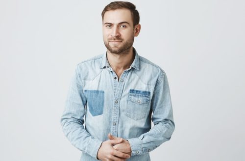 Portrait of puzzled european male wears fashionable denim clothes, keeps hands in together, raises eyebrow in bewilderment, has to make difficult life choice, looks confused